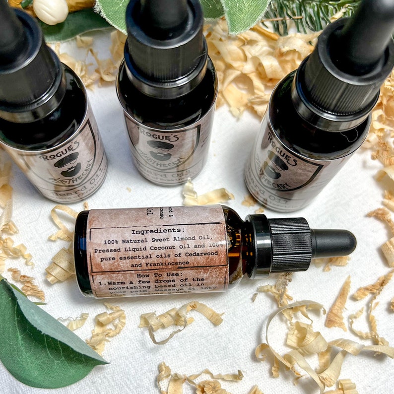 K&ouml;nnte beinhalten: Drei kleine braune Glasflaschen mit schwarzen Tropfern und Etiketten mit der Aufschrift "Rogue's Apothecary Beard Oil". Die Flaschen stehen auf einer Oberfl&auml;che, die mit Holzsp&auml;nen bedeckt ist. Das Etikett auf einer Flasche listet die Zutaten und Gebrauchsanweisungen auf.