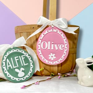 May include: Two personalised Easter egg-shaped tags with white lace trim. The green tag says "Alfie" with a white bunny rabbit silhouette. The pink tag says "Olive" with a white flower.