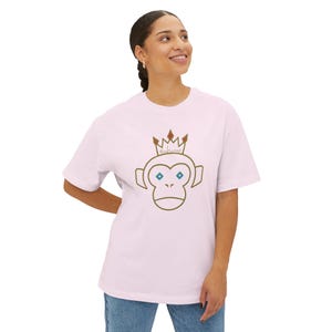 May include: Light pink t-shirt featuring a cartoon monkey wearing a crown. The monkey has blue eyes and the text "Girl Sense" is above the monkey's head. The shirt is being worn by a person.