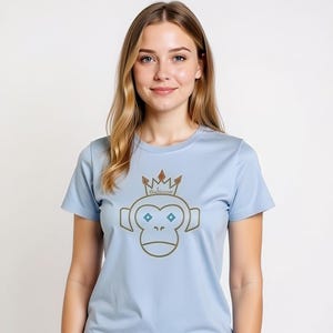 May include: Light blue t-shirt featuring a cartoon monkey wearing a crown. The monkey has brown outlines, blue diamond-shaped eyes, and a sad expression. The crown has brown and red accents, and the text "Guillaume" is above the monkey's head.