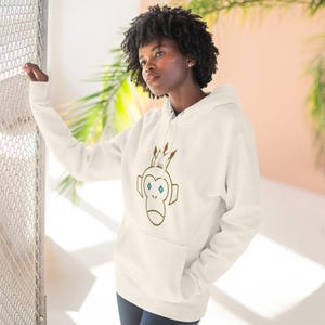 May include: Cream-colored hoodie featuring a cartoon monkey wearing a crown. The monkey has blue eyes and is outlined in brown. The hoodie has a front pocket and a drawstring hood. The background is a mix of pink and green.