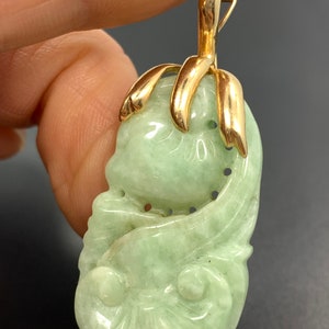 May include: A green jade pendant carved with a floral design, set in a gold tone metal frame. The pendant is attached to a gold tone metal bail.