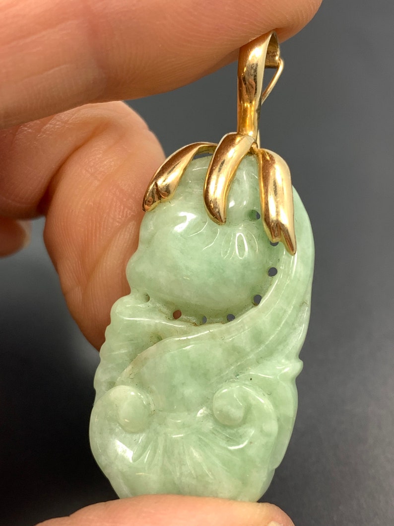 May include: A green jade pendant carved with a floral design, set in a gold tone metal frame. The pendant is attached to a gold tone metal bail.