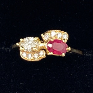 May include: A gold ring with a ruby and diamond setting. The ruby is oval shaped and the diamonds are set in a pave setting around the ruby. The ring is on a black background.