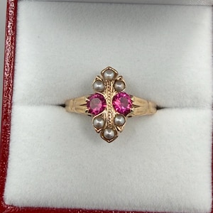 May include: A gold ring with two large pink gemstones and small pearls. The ring is set in a decorative pattern and is displayed in a red-lined jewelry box. The ring's design is symmetrical, with the gemstones and pearls arranged in a balanced composition.
