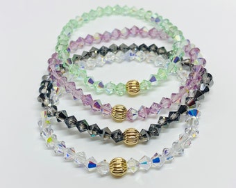 May include: Four crystal bracelets with gold accents. The bracelets are stacked on top of each other and are in the colors clear, light green, light purple, and dark gray.