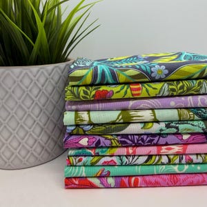 May include: A stack of colorful fabric squares with floral and botanical patterns. The fabrics feature a variety of colors, including green, purple, pink, and blue. A gray ceramic pot with a green plant is in the background.