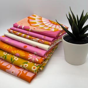 May include: A stack of eight fabric squares in pink, orange, and yellow with floral and geometric patterns. The fabric squares are folded and stacked on a white surface.