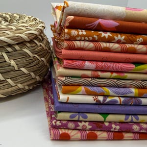 May include: A stack of folded fabric squares featuring various floral and geometric patterns in shades of orange, pink, purple, and cream, positioned next to a round, woven, light brown straw basket with a textured, braided design.