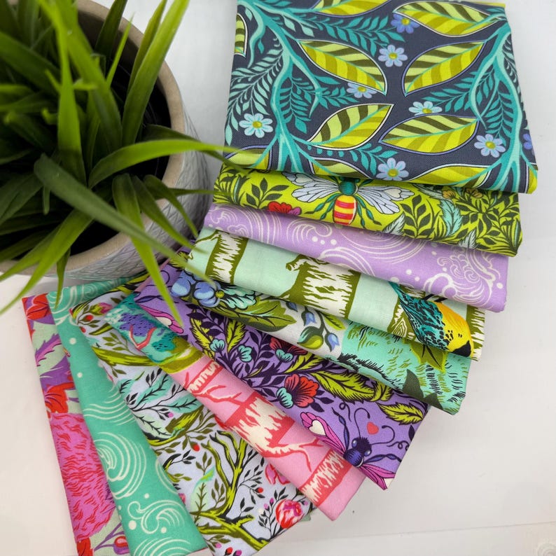 May include: A stack of colorful fabric squares with various floral and insect patterns. The fabrics feature shades of green, purple, pink, and blue. A potted plant is in the background.