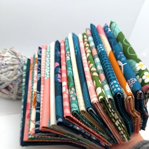 May include: A stack of folded fabric squares in various colors and patterns. The fabrics feature floral and geometric designs in shades of teal, pink, green, and white. A ball of yarn is visible in the background.