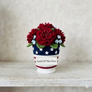 May include: A crocheted floral arrangement in a patriotic theme. The arrangement features vibrant red flowers, small blue accents, and green leaves, set in a vase with a navy blue base adorned with white stars and red and white stripes. The text "Land Of The Free" is visible.