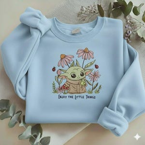 Op de afbeelding: Lichtblauw sweatshirt met een geborduurd ontwerp van een stripfiguur, roze bloemen, lieveheersbeestjes en de tekst "ENJOY THE LITTLE THINGS". De mouwen zijn opgerold.