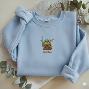 May include: A light blue sweatshirt featuring an embroidered image of a green alien character holding a sword, with the word "Huzzah" below. The sleeves are cuffed, and the overall design is simple and clean.