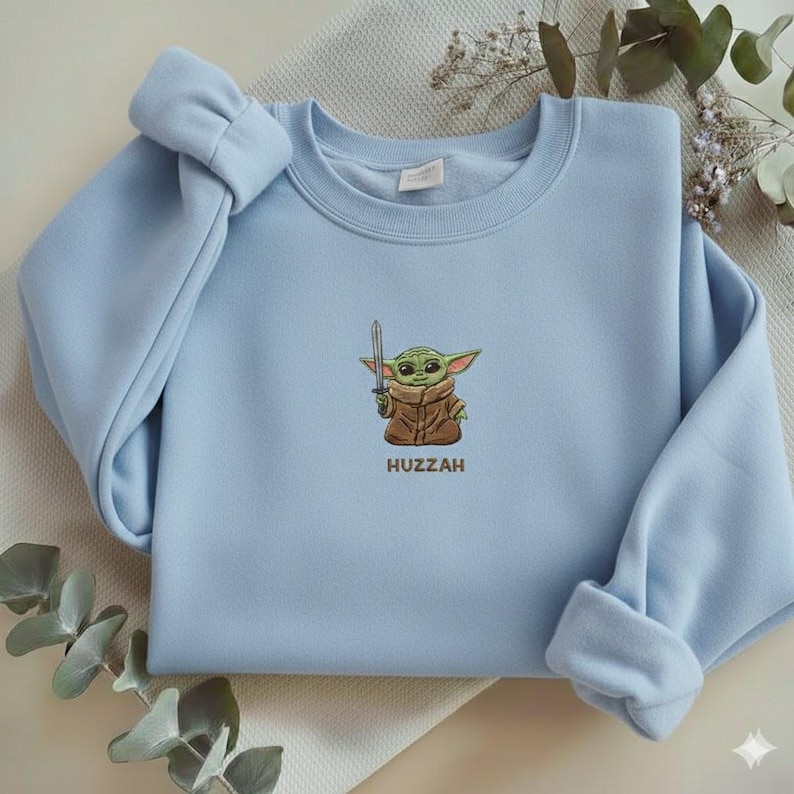 May include: A light blue sweatshirt featuring an embroidered image of a green alien character holding a sword, with the word "Huzzah" below. The sleeves are cuffed, and the overall design is simple and clean.
