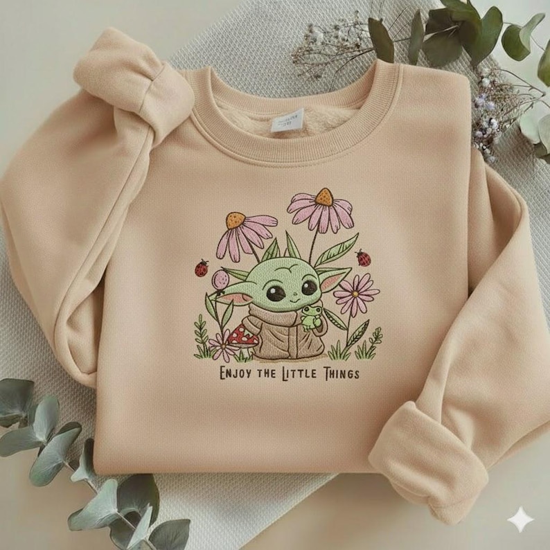 Op de afbeelding: Beige sweatshirt met een geborduurd ontwerp van een groen alien-personage, roze bloemen, lieveheersbeestjes en de tekst "Enjoy the Little Things". De sweatshirt heeft lange mouwen en een ronde hals.
