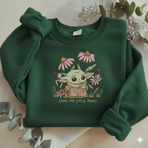 Op de afbeelding: Donkergroene sweatshirt met een geborduurd ontwerp met een stripfiguur omringd door roze bloemen en de tekst "Enjoy the Little Things". Het ontwerp bevat een kleine kikker en lieveheersbeestjes.
