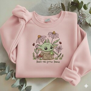 Op de afbeelding: Een lichtroze sweatshirt met een geborduurd ontwerp van een groen, buitenaards personage dat een kleine groene kikker vasthoudt, omringd door bloemen en lieveheersbeestjes. De tekst "ENJOY THE LITTLE THINGS" staat eronder.
