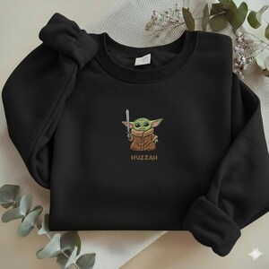 May include: Black crewneck sweatshirt featuring an embroidered image of a green alien character holding a silver sword. The character is wearing a brown coat. The word "Huzzah" is embroidered below the character.