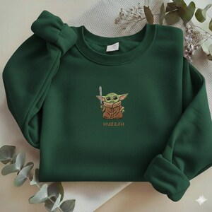 May include: Dark green sweatshirt featuring an embroidered image of a green alien character holding a silver sword. The character is wearing a brown coat. The word "Huzzah" is embroidered below the character.