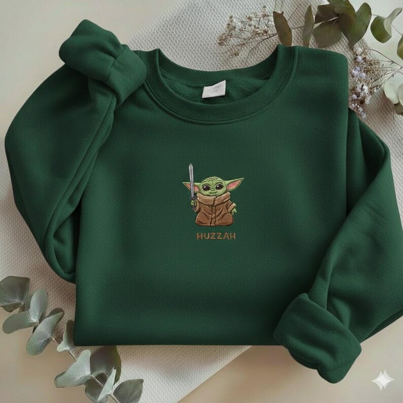 May include: Dark green sweatshirt featuring an embroidered image of a green alien character holding a silver sword. The character is wearing a brown coat. The word "Huzzah" is embroidered below the character.