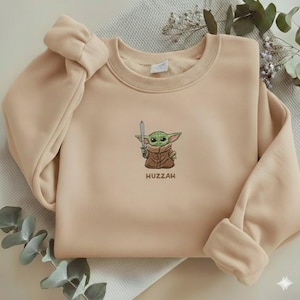 May include: A beige sweatshirt featuring an embroidered image of a green alien character holding a sword. The text "Huzzah" is below the character. The sweatshirt has a crew neck and long sleeves with cuffed wrists.