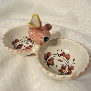 May include: A white ceramic divided dish with a bird-shaped handle. The dish features red and yellow floral designs and a pink dotted border. The bird is pink and white with a yellow beak and blue eye.
