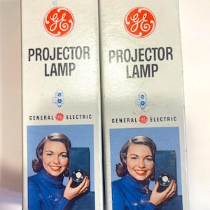 May include: Two vintage General Electric Projector Lamp boxes. Each box features the GE logo, the words "PROJECTOR LAMP", and a retro image of a woman holding a projector. The boxes are marked "Made in U.S.A."