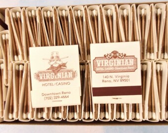 May include: A box of vintage matchbooks from the Virginian Hotel/Casino in Reno, Nevada. The matchbooks are beige with brown text and graphics, including a cowboy illustration. The matches are brown with white tips.