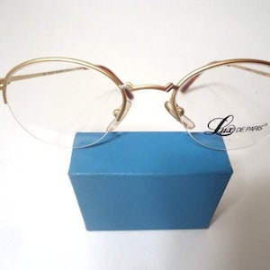 May include: Gold-rimmed spectacles with clear lenses, resting on a blue box. The glasses have a delicate, oval shape and the brand name "Lux de PARIS" is printed on the lens. The frame is a warm gold colour.