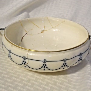 Puede incluir: Cuenco de cerámica color crema con borde dorado y ribete decorativo azul. El cuenco tiene dos asas y está agrietado en varios lugares. El ribete azul presenta un patrón repetitivo de círculos y formas de lágrima.