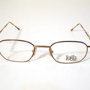 May include: Oval-shaped eyeglasses with a thin, brown metal frame and clear lenses. The temples have a patterned design. The brand name "Logo" is printed on the lower right lens.