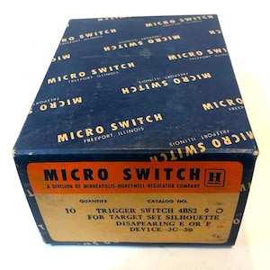 May include: A vintage Micro Switch box in navy blue with gold lettering. The box features the text "MICRO SWITCH FREEPORT ILLINOIS" repeated across the top. The front label reads "MICRO SWITCH" and includes the text "TRIGGER SWITCH 4BS2" and other technical details.