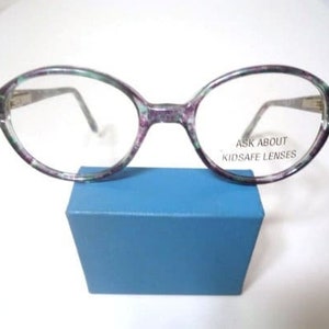May include: Oval-shaped eyeglasses with a speckled purple and green frame, resting on a blue box. The lenses are clear, and the text "ASK ABOUT KIDSAFE LENSES" is visible on one lens. The temples are silver.