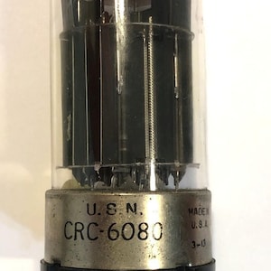 Puede incluir: Válvula de vacío vintage con una cubierta de vidrio transparente y una base de metal. La base está marcada con "U.S.N. CRC-6080" y "MADE IN U.S.A." El tubo tiene componentes internos y pines.