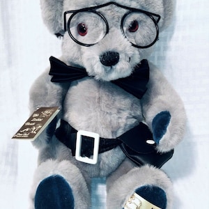 May include: A gray plush bear wearing black-rimmed glasses, a black bow tie, and a black belt with a white buckle. The bear has dark blue accents on its paws and ears. A tag reads "Aries".