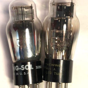 Puede incluir: Dos válvulas de vacío vintage con bombillas de vidrio transparente y bases negras. Una válvula está etiquetada como "NG-SOL" y "IN U. S. A.", la otra como "RENEWING" y "ELCO TUBE". Ambas tienen múltiples pines.