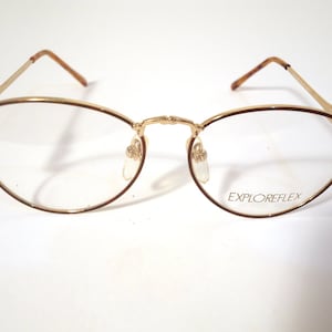 Könnte beinhalten: Goldfarbene Brille mit ovalen Gläsern und dünnen Rahmen. Die Ränder sind dunkelbraun und die Bügel haben ein Schildpattmuster. Der Text "EXPLOREFLEX" ist auf dem unteren rechten Glas aufgedruckt.