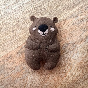 Peut inclure: Un animal en peluche en feutre brun en forme d'ours avec un nez noir et un sourire cousu. L'ours a un nez et des joues brun clair.