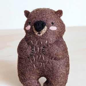 Peut inclure: Un animal en peluche en feutre marron en forme de wombat. Le wombat a un nez noir, des joues roses et un sourire cousu.
