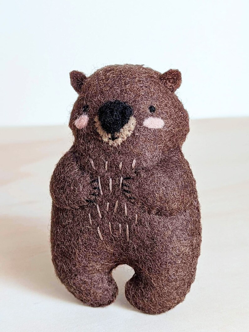 Peut inclure: Un animal en peluche en feutre marron en forme de wombat. Le wombat a un nez noir, des joues roses et un sourire cousu.