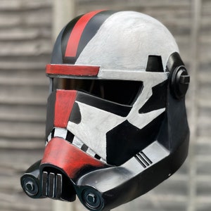 May include: A black, red, and white helmet with a visor. The helmet has a red stripe on top and red accents on the front. The design features a mix of black, white, and red paint, creating a striking visual effect. The helmet is likely for cosplay or display.