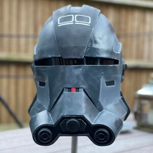May include: A dark gray helmet with a matte finish, featuring a rectangular visor and a series of geometric shapes. The helmet is mounted on a silver stand. The top of the helmet has two white rectangles. A red accent is visible near the center.