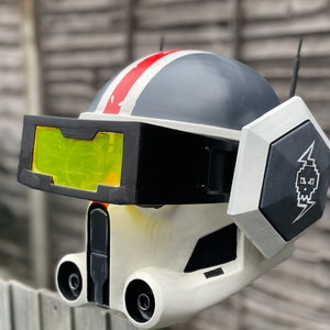 May include: A detailed, off-white and gray helmet with a black visor and a red stripe. The helmet features a black and white emblem with a skull and lightning bolt. The design includes two small antennas and a yellow-tinted visor.