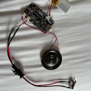 Puede incluir: Un conjunto de componentes electrónicos, que incluyen una placa de circuito negro etiquetada "GC03", un pequeño altavoz, una batería y varios cables con conectores. Los componentes están conectados con cables rojos y negros.