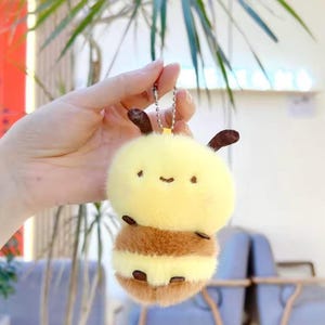 May include: A yellow and brown plush keychain shaped like a cartoon bee. The keychain has a yellow head with small black eyes and a smiling mouth. The body is brown and yellow striped. The keychain is attached to a silver chain.