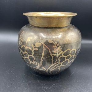May include: A round, dark metal pot with a brass rim and a brass interior. The pot features a floral design etched into the metal. The pot is likely used for decorative purposes.