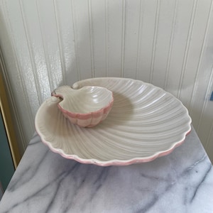 May include: A white ceramic seashell-shaped serving dish with a smaller, matching dip bowl. The dish has a pink trim and a textured surface. The dip bowl is also white with a pink trim. The set is designed to resemble a seashell.