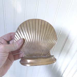 May include: A golden-colored seashell-shaped bookend. The bookend has a fan-like design with vertical lines radiating from the base. The base is a rounded rectangle. The bookend is made of metal.