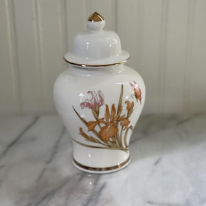 May include: A white ceramic jar with a lid, featuring a floral design of orange and pink irises and a gold trim. The jar has a rounded shape and a small gold finial on the lid. The design is painted on the white surface.
